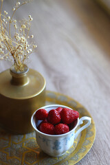 Porcelain cup filled with fresh strawberries, golden vase with gypsophila flowers and vintage tray on the table. Selective focus.