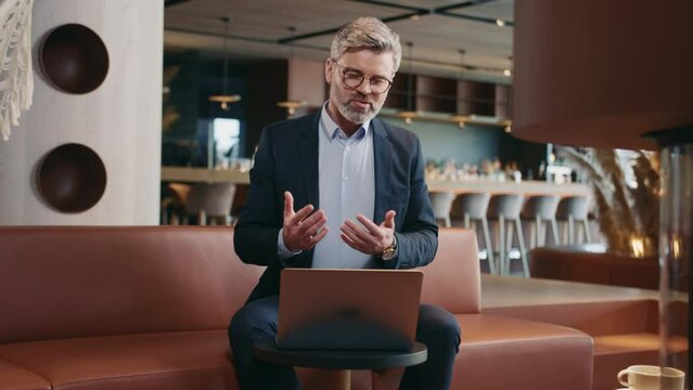 Serious confident Caucasian gray-haired man sitting at laptop, speaking to webinar listeners. Portrait of handsome middle-aged experienced business trainer working online. Daytime, computer