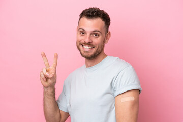 Young Brazilian man wearing a band-aids isolated on pink background smiling and showing victory sign