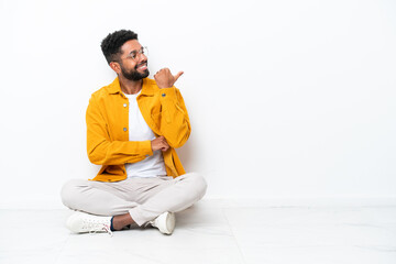 Young Brazilian man sitting on the floor isolated on white background pointing to the side to...