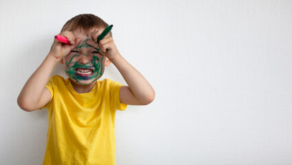 A 4-year-old boy painted his face with a marker. The boy stands against a white wall, holding markers in his hands and portrays a monster with horns. Banner with space for text