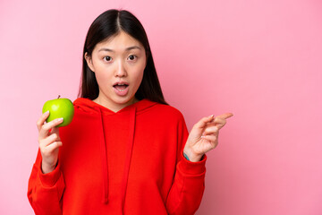 Young Chinese woman with an apple isolated on pink background surprised and pointing side