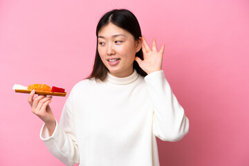 Young Chinese woman holding sashimi isolated on pink background listening to something by putting hand on the ear