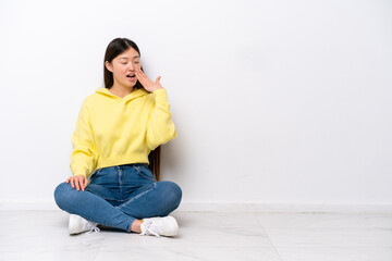 Young Chinese woman sitting on the floor isolated on white wall yawning and covering wide open mouth with hand