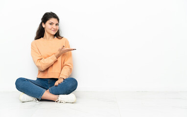 Teenager Russian girl sitting on the floor presenting an idea while looking smiling towards