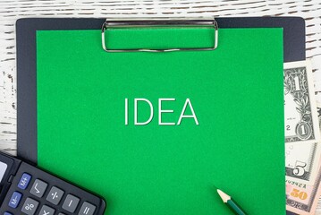 IDEA - word (text) dollar bills on green background, pencil, calculator and wooden white table. Business concept: buying, selling, commerce (copy space).