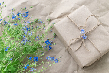 On a craft background, a gift in craft packaging with a bow made of a tourniquet next to blue wild flowers. The gift is made by hand. Eco-friendly packaging. Ecology. Layout
