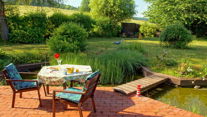 romantischer Frühstücksplatz am Gartenteich mit Holzbrücke neben roter Terrasse und grünem...