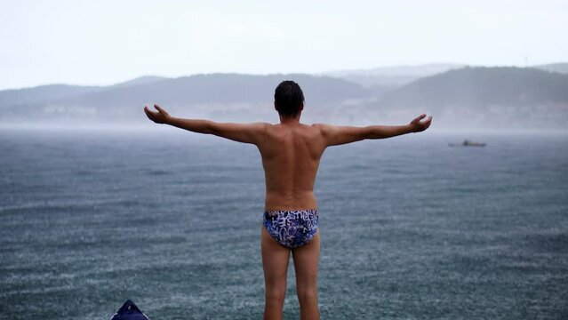 Happy adult male swimmer standing in the pouring rain, arms wide open, by the ocean after a swim.