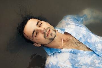 A man with closed eyes with dark long hair lies in the water, top view