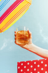 Human hand holding glass of whiskey over summer blue sky background. Vacation, happiness, summer...