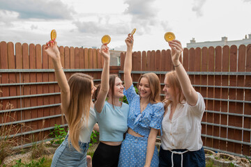 Laughing young girls standing together, raising hands with oranges and looking at each other, sky...
