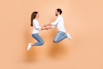 Fototapeta na wymiar Full length body size view of two attractive cheery partners jumping holding hand isolated over beige pastel color background