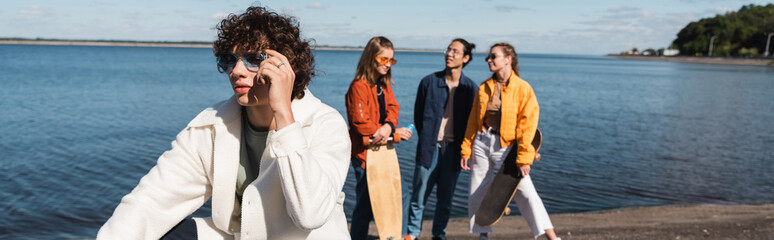 curly man in trendy sunglasses near multiethnic skaters talking by river, banner.