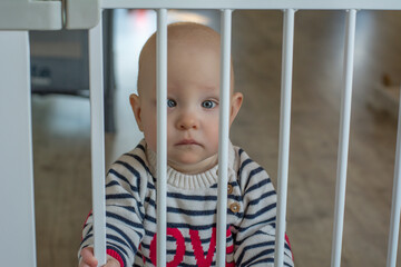 The little sad baby with blue eyes is standing behind the bars. The bars in the room against little...