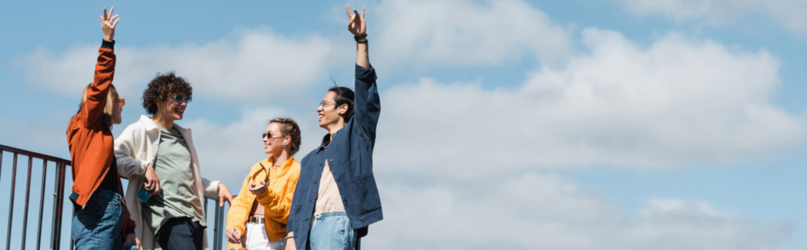 Happy Asian Man Showing Rock Sign Near Friends Against Blue Cloudy Sky, Banner.