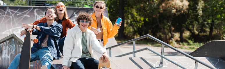cheerful and stylish multicultural skaters looking at camera in skate park, banner.