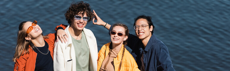 joyful asian man showing victory sign near river and trendy friends, banner.