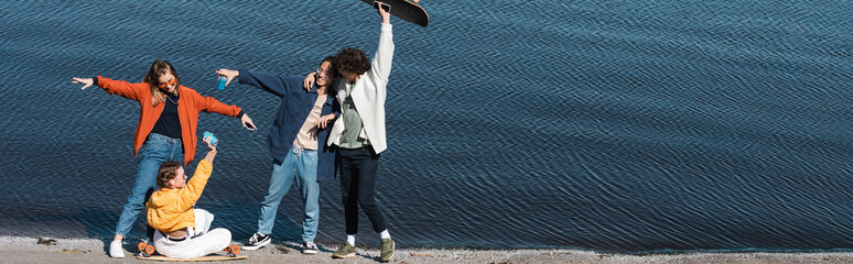 happy multiethnic skaters in stylish clothes having fun near river, banner.