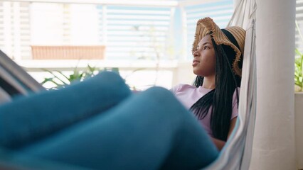African american woman covering face with hat lying on hammock at home terrace