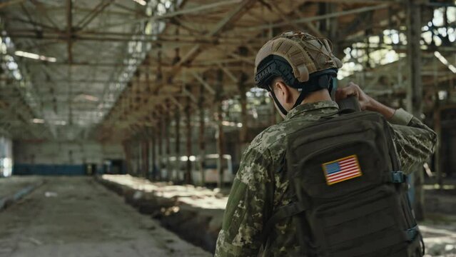 Back view of strong american fighter in military uniform and helmet carrying backpack while walking alone on abandoned plant. Concept of army, mission and bravery.