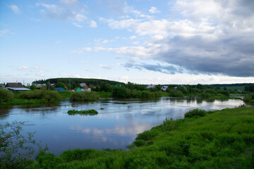 a village on the river bank with bright green grass and a beautiful sky.