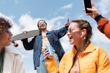 low angle view of asian man with longboard showing victory sign near happy friends.