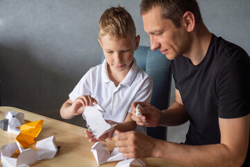 A cute boy is sitting with his dad at the table and collecting origami. Glue the parts