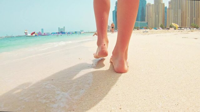 Legs feet of young female walking sand barefoot on the beach. Slim female legs and feet walking along sea water waves on sandy beach. Pretty woman walks at seaside surf. Summer vacation in ocean.