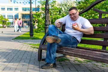 Fototapeta premium A cheerful young man, sitting on a bench with a donut, spilled coffee on his jeans. 