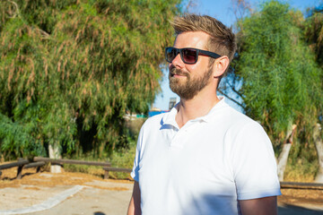 Outdoor shot of beautiful bearded young man in sunglasses posing over city garden on warm sunny...