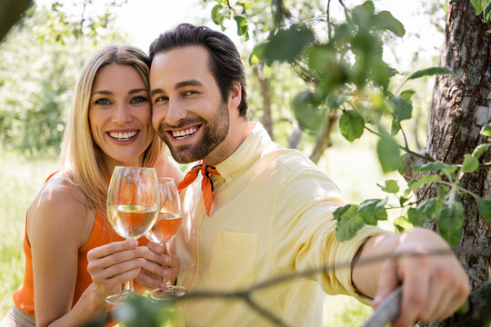 Smiling and trendy couple holding glasses of wine near tree in summer park.