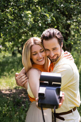 Stylish couple embracing while taking photo on retro camera in park.