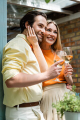 Stylish woman touching face of smiling boyfriend with wine in outdoor cafe.