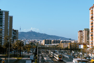 Naklejka premium Barcelona, Spain - April 16, 2022 : View of a train station and tall apartment buildings in the neighborhood of El Gornal Bellvitge. Tibidabo, church or temple on a hill in the distance