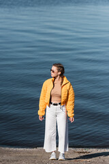 full length of woman in white trousers and yellow jacket looking away on river bank.