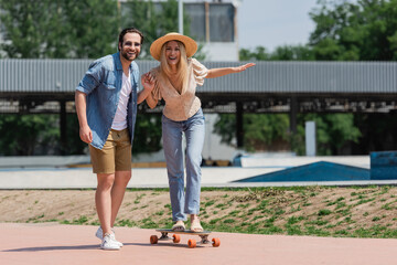 Positive couple holding hands and looking at camera while riding longboard in skate park.