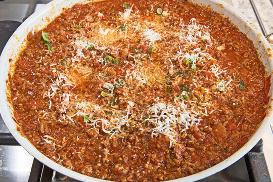 A delicious pan of Bolognese sauce, sprinkled with melting cheese and basil leaves, sits on top of a stove and is ready for serving.