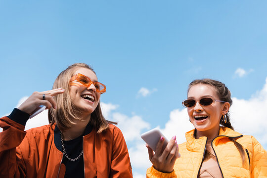 woman in stylish sunglasses holding smartphone near happy friend against blue sky.