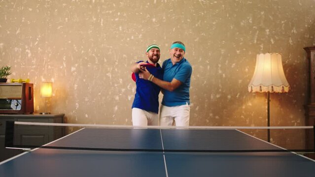 Good looking one man old looking and one young man playing together like a team in the ping pong they win the game get excited and very happy hugging each other while holding the racket