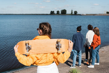 back view of young woman with skateboard standing near river and blurred friends.