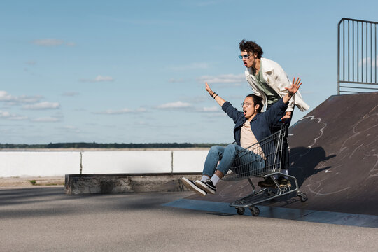 excited interracial friends moving on shopping cart down from skate ramp.