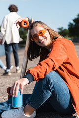 cheerful woman in sunglasses looking at camera near skateboard and soda can.
