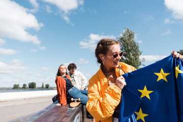 woman in trendy sunglasses smiling while holding eu flag near blurred friends. - obrazy, fototapety, plakaty