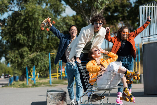 young and cheerful multiethnic skaters having fun with shopping cart in park.