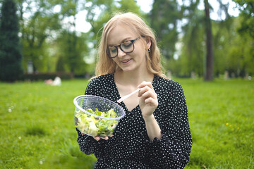 Happy European 30s woman in dress sitting on grass and eating yummy salad from plastic lunch box...