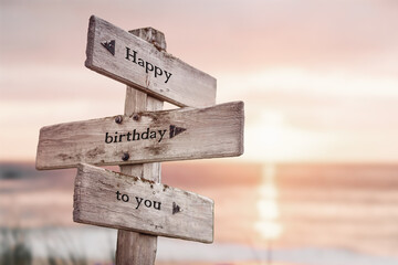 happy birthday to you text quote on wooden crossroad signpost outdoors on beach with pink pastel...
