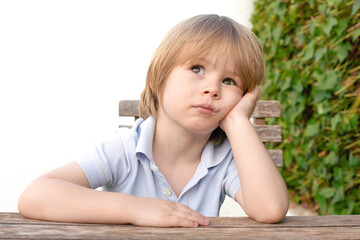 Naklejka premium Cute blond haired boy sitting at the table and doubtful .Fresh green leafs on the background.