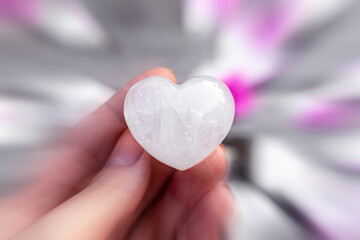 Heart-shaped clear quartz crystal in a female hand, pink rose petals background, radial blur