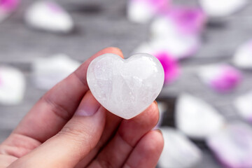Heart-shaped clear quartz crystal in a female hand, pink rose petals background - obrazy, fototapety, plakaty